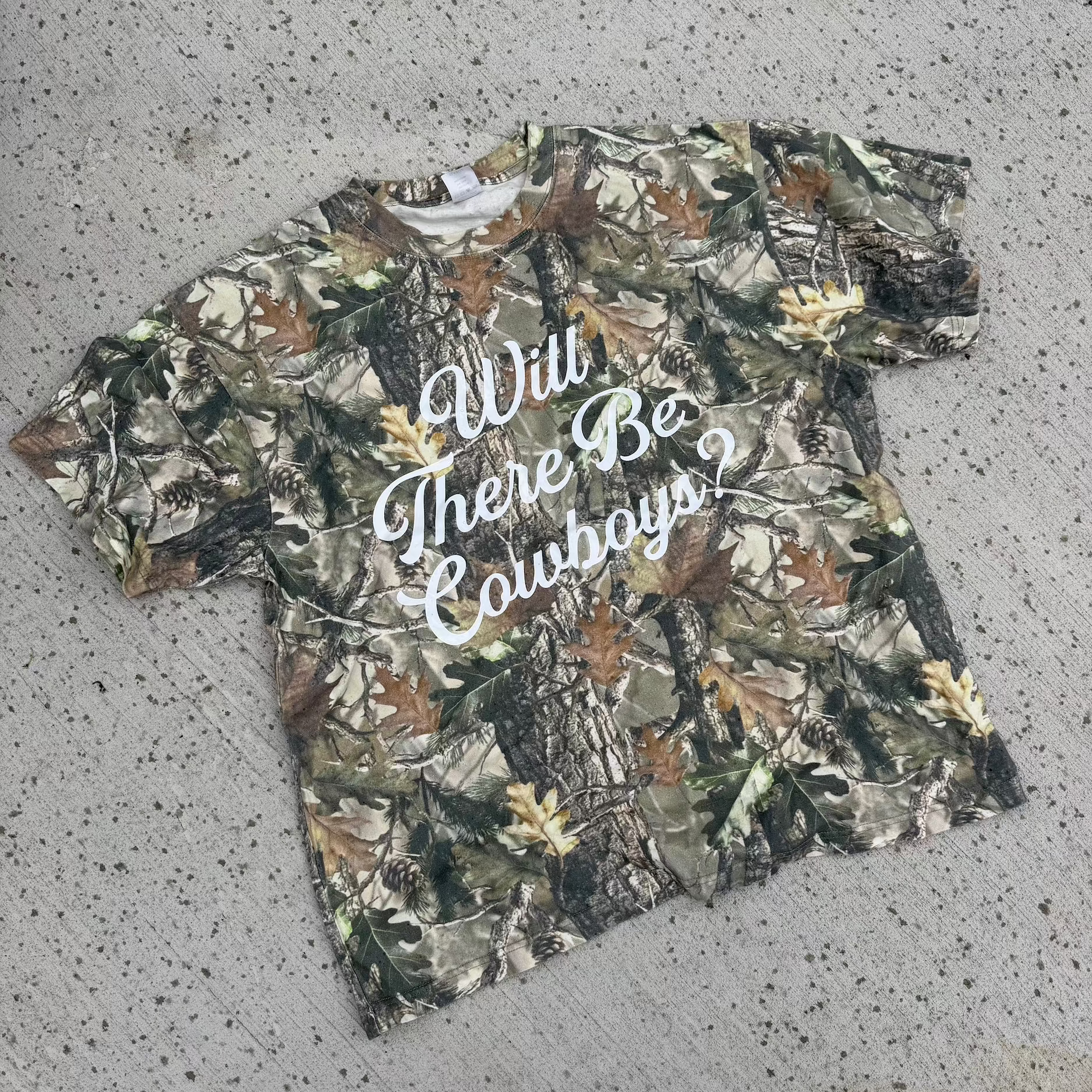 Camouflage t-shirt with 'Will There Be Cowboys?' text on a concrete surface